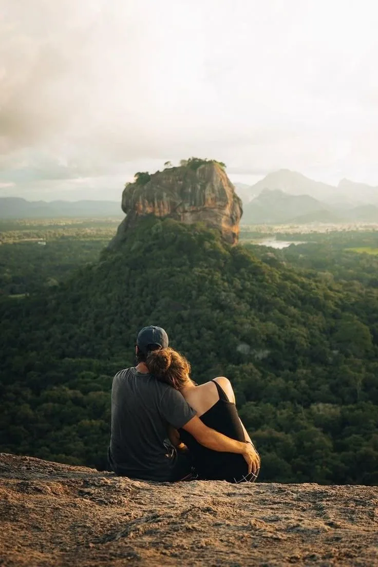 Two travelers sit together above the forest with Sigiriya Rock in the distance.